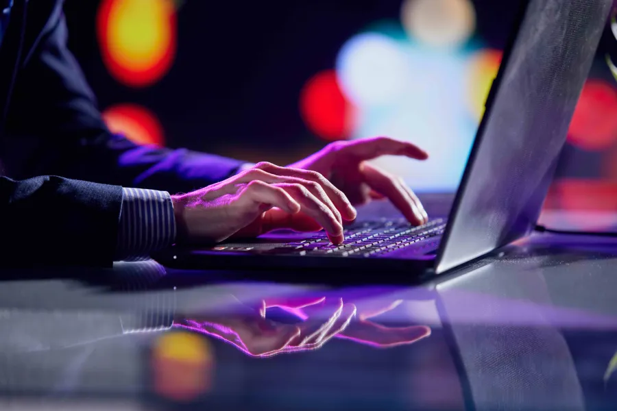 Person typing on a laptop keyboard with colorful blurred lights in the background and reflections on the glass surface.